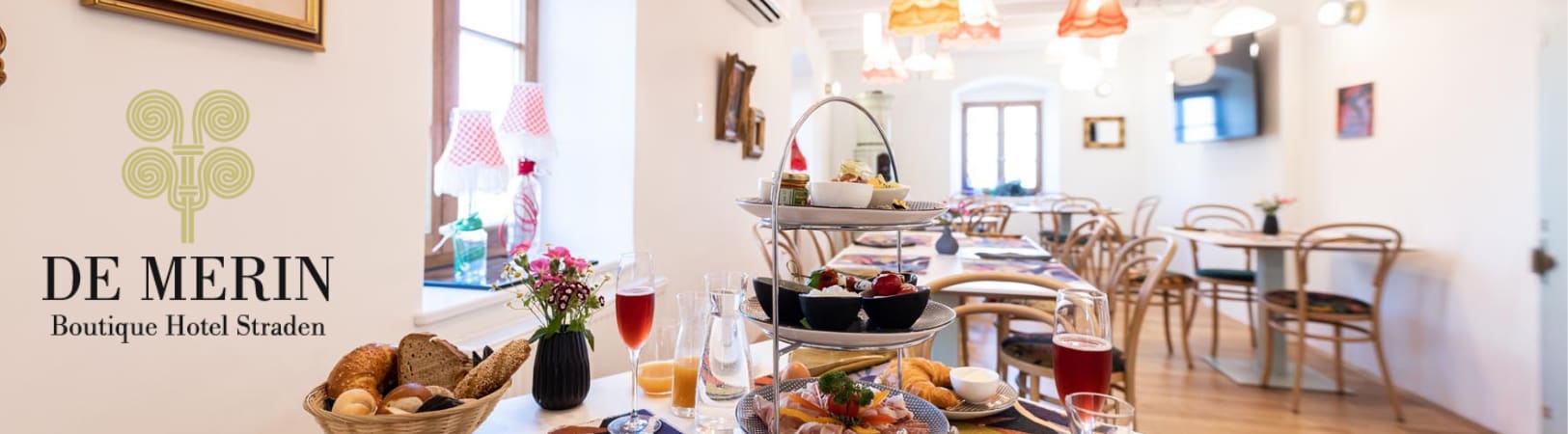 ALT-Tag: Eleganter Speisesaal in einem Boutique-Hotel mit Holzböden und hellen Wänden. Auf einem Tisch im Vordergrund stehen ein Frühstücksarrangement mit Brot, Aufschnitt, frischen Säften und einem dreistufigen Servierständer mit verschiedenen Speisen. Im Hintergrund sind weitere Tische mit Stühlen und Lampenschirmen in warmen Farbtönen zu sehen. Links ist das Logo des Hotels "De Merin Boutique Hotel Straden" in Schwarz und Gold dargestellt.