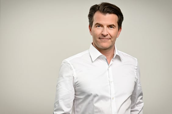 Ein Mann trägt ein klassisches weißes Hemd mit aufgekrempelten Ärmeln. Der Hintergrund ist neutral hellgrau. Sein Haar ist ordentlich gestylt.