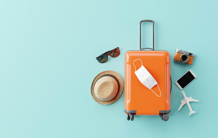 Ein oranges Handgepäck auf einem hellblauen Hintergrund, darauf liegt eine weiße Gesichtsmaske. Daneben befinden sich eine Sonnenbrille, ein Strohhut, eine Kamera, ein Mobiltelefon und ein kleines Modellflugzeug.