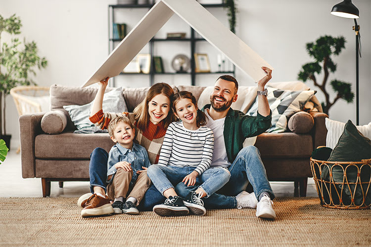 Eine Familie sitzt gemeinsam auf einem Teppich in einem gemütlichen Wohnzimmer vor einem braunen Sofa. Im Hintergrund sind Bücherregale und einige Pflanzen zu sehen. Die Eltern sitzen hinten und halten ein weißes, dreieckiges Brett über ihren Köpfen, das wie ein stilisiertes Hausdach aussieht. Vor ihnen sitzen zwei Kinder. Die Szene strahlt eine harmonische und warme Atmosphäre aus.