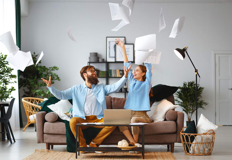 Ein Mann und eine Frau sitzen auf einem Sofa in einem modernen Wohnzimmer und werfen freudig zahlreiche Blätter Papier in die Luft. Vor ihnen steht ein Kaffeetisch mit einem geöffneten Laptop darauf. Die Umgebung ist hell und geschmackvoll dekoriert, mit Pflanzen, beleuchteter Stehlampe und Regalen im Hintergrund.