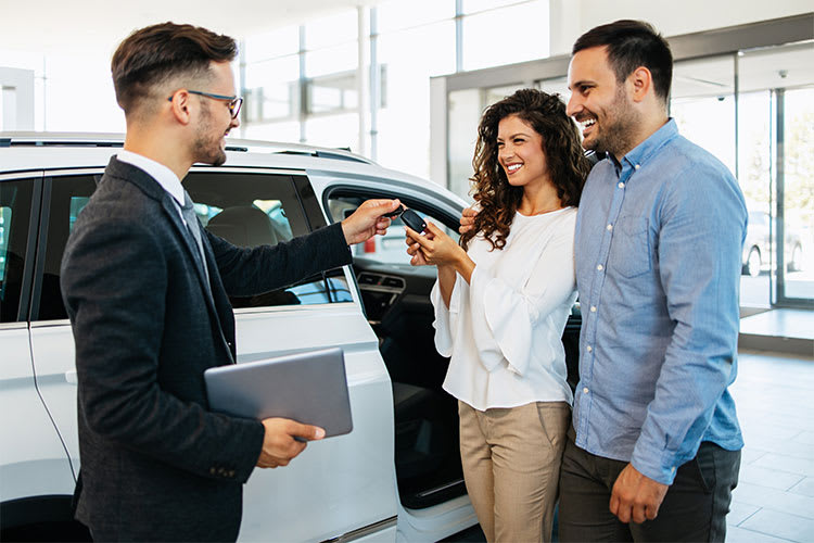 Ein Autoverkäufer in einem Anzug übergibt einem jungen Paar den Autoschlüssel. Das Paar steht vor einem weißen Auto in einem modernen Autohaus. Der Mann hält ein Tablet in der Hand. Die Gesichter der Personen sind unscharf.