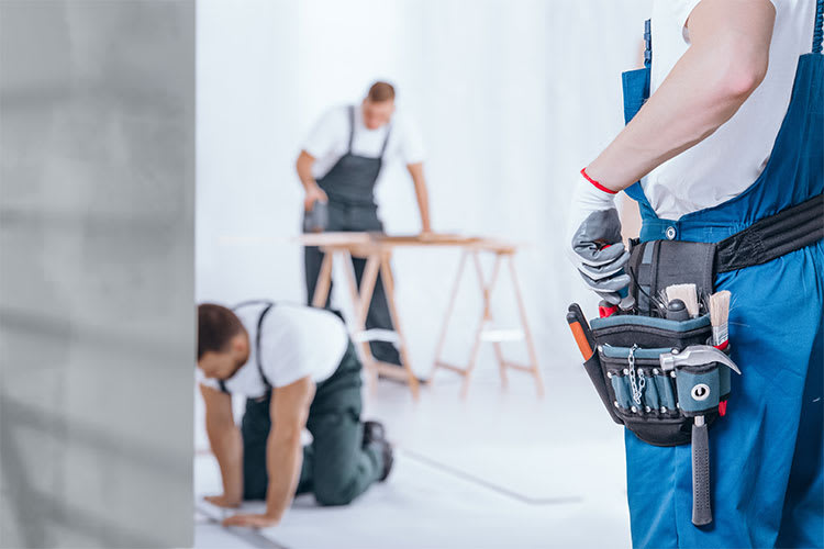 Drei Handwerker in blauen Latzhosen und weißen T-Shirts arbeiten in einem hellen Raum. Im Vordergrund steht ein Arbeiter mit einem gut gefüllten Werkzeuggürtel, der Schraubendreher, Zangen und andere Werkzeuge enthält. Im Hintergrund kniet ein Arbeiter auf dem Boden, während ein weiterer an einem Tisch arbeitet. Alle tragen Arbeitshandschuhe.