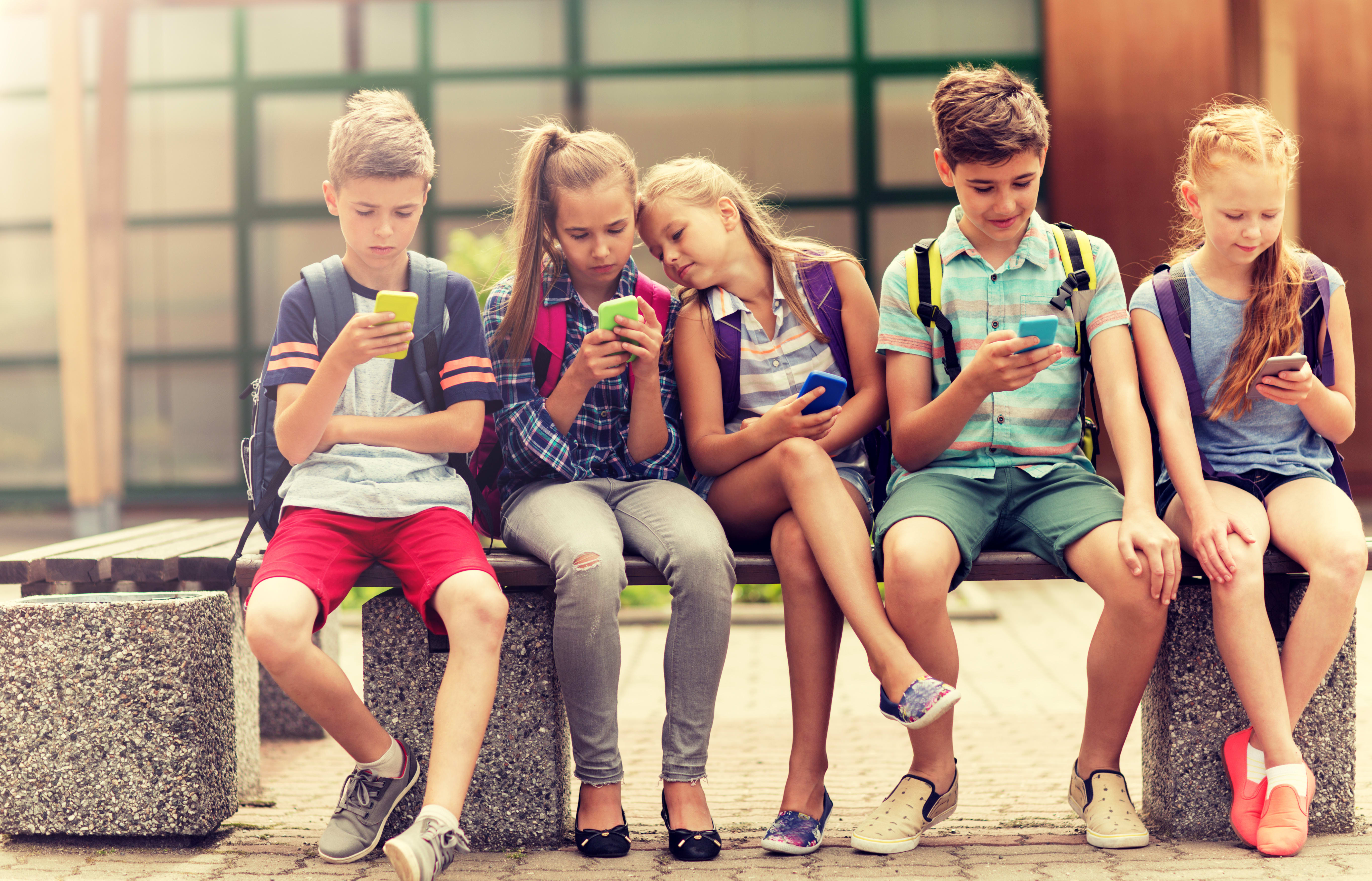Fünf Kinder im Teenageralter sitzen auf einer Bank im Freien. Sie tragen Sommerkleidung und Rucksäcke und konzentrieren sich auf ihre Smartphones. Die Gesichter der Kinder sind unscharf.