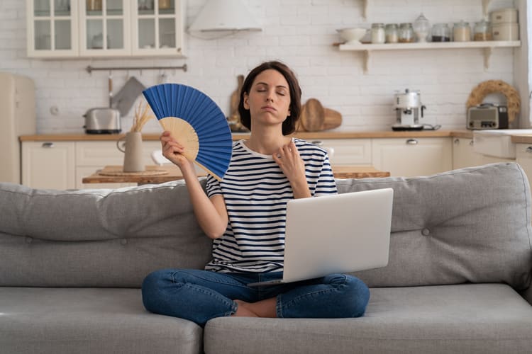 Eine Person sitzt auf einem grauen Sofa in einer modernen Küche. Sie trägt ein gestreiftes T-Shirt und Jeans. In einer Hand hält sie einen blauen Fächer, mit dem sie sich Luft zufächelt, während auf ihrem Schoß ein aufgeklappter Laptop ruht. Hinter ihr sind weiße Küchenschränke, offene Regale mit Geschirr und eine Arbeitsplatte mit verschiedenen Küchengeräten zu sehen.