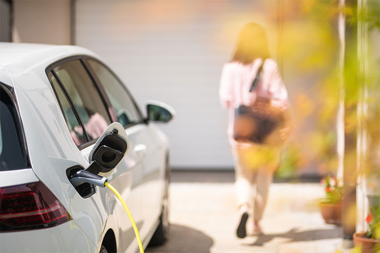 Ein weißes Elektroauto wird über ein gelbes Kabel aufgeladen. Im Hintergrund geht eine unscharf abgebildete Frau in einem hellen Outfit und mit einer Tasche in der Hand auf ein Gebäude zu. Es ist ein sonniger Tag.