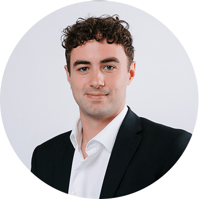 Ein Mann in einem schwarzen Anzug mit weißem Hemd, davor ein neutraler weißer Hintergrund. Das Gesicht ist unscharf dargestellt. Der Mann hat lockiges, braunes Haar.