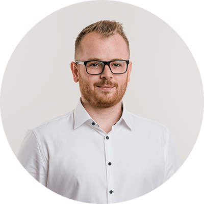 Eine Person mit blonder Kurzhaarfrisur, die ein weißes Hemd mit schwarzen Knöpfen trägt, vor einem einfarbigen, hellbeigen Hintergrund.