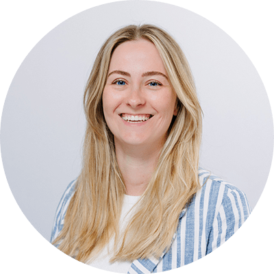 Eine Person mit langen, glatten, hellblonden Haaren, die ein helles, gestreiftes, blau-weißes Hemd über einem weißen Oberteil trägt, vor einem neutralen, hellgrauen Hintergrund.