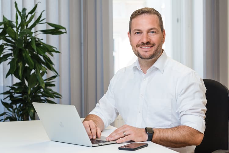 Ein Mann in einem weißen, langärmligen Hemd sitzt an einem Bürotisch. Vor ihm steht ein aufgeklappter Laptop, und eine Smartwatch ist an seinem linken Handgelenk zu sehen. Am Tisch liegt ein Smartphone. Im Hintergrund befindet sich eine große grüne Pflanze, und Vorhänge sowie ein Fenster sind sichtbar.