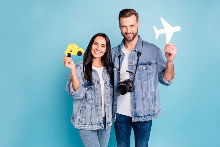 Zwei Personen in Jeansjacken stehen vor einem hellblauen Hintergrund. Beide halten Papiersymbole: Die linke Person hält ein gelbes Taxi und die rechte Person ein weißes Flugzeug, während eine Kamera um ihren Hals hängt. Sie tragen lässige Kleidung, die Kombination von Jeans und weißen T-Shirts.