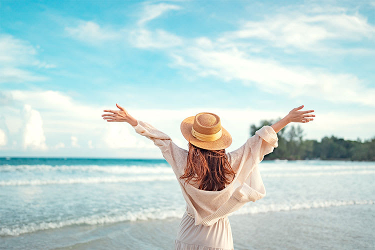 Frau mit langen, braunen Haaren steht am Strand, trägt einen Strohhut und eine weiße Bluse. Sie breitet ihre Arme weit aus, als würde sie die frische Meeresbrise und die Freiheit des offenen Himmels genießen. Im Hintergrund sind das Meer und ein bewaldetes Ufer zu sehen. Der Himmel ist blau mit einigen weißen Wolken.