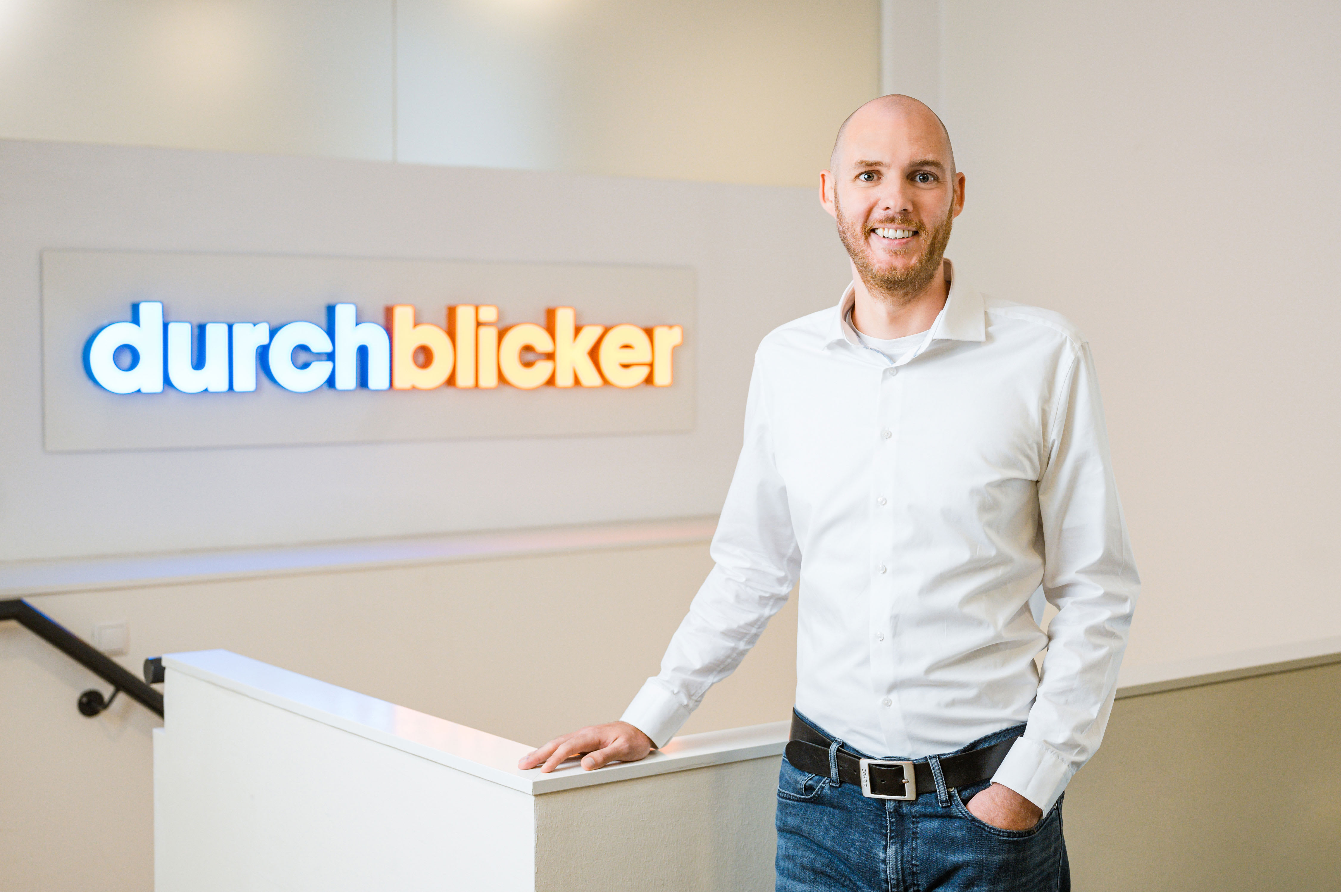 Ein Mann in einem weißen Hemd und blauen Jeans steht auf einer Treppe. Im Hintergrund ist eine Wand mit einem beleuchteten Logo der Aufschrift "durchblicker" in blauen und orangefarbenen Buchstaben.