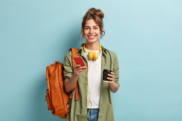 Eine Person steht vor einem hellblauen Hintergrund. Sie trägt einen orangefarbenen Rucksack über einer Schulter und hält in einer Hand ein rotes Smartphone sowie in der anderen Hand einen schwarzen Kaffeebecher. Die Person trägt ein olivgrünes Hemd offen über einem weißen T-Shirt und hat gelbe Kopfhörer um den Hals hängen. Die Haare sind zu einem hohen, lockeren Knoten gebunden.