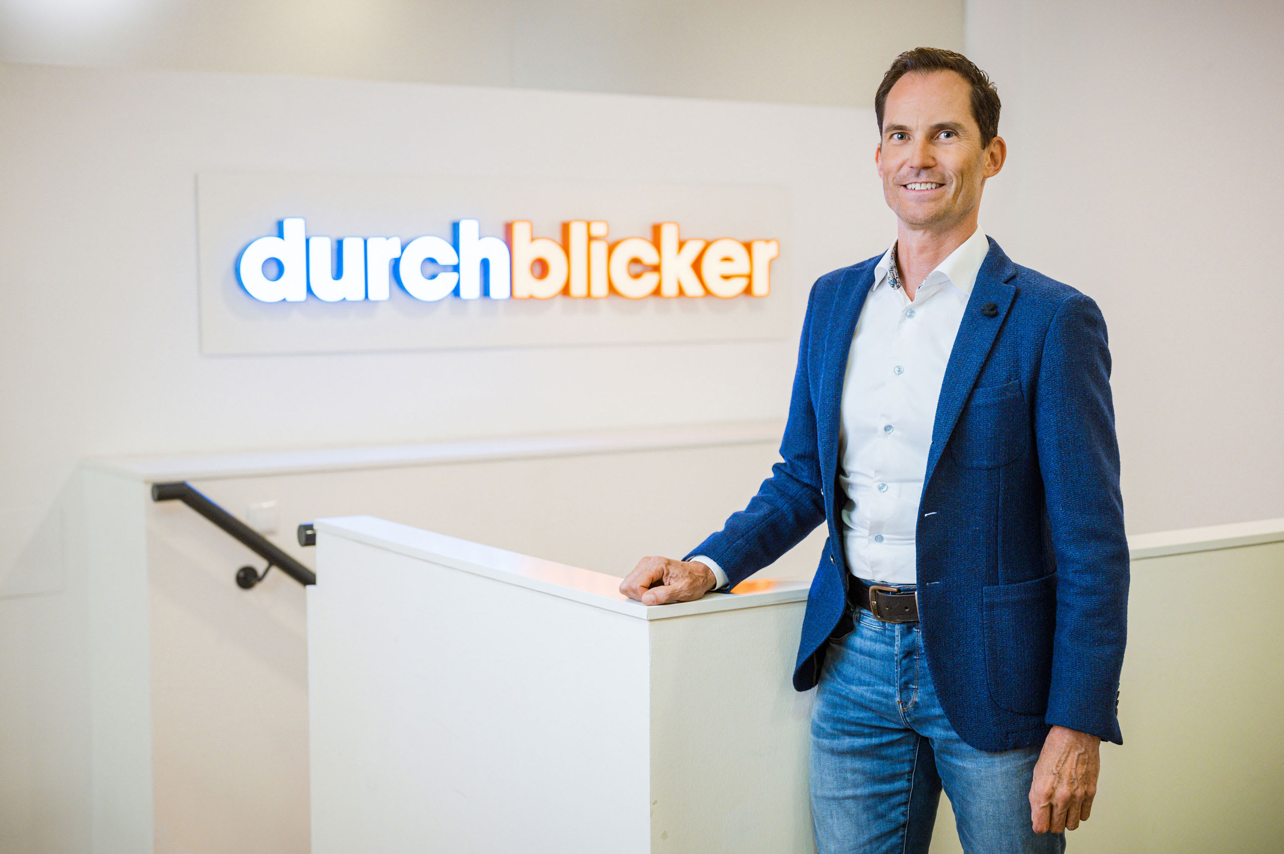 Ein Mann in einem blauen Sakko und einer Jeans steht an einem weißen Geländer. Im Hintergrund ist ein beleuchtetes Schild mit dem Schriftzug "durchblicker" zu sehen. Der Raum hat eine moderne Einrichtung.