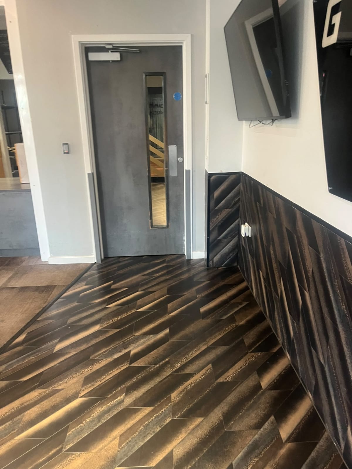 Grey interior door with narrow vision panel, wall-mounted TV, and dark geometric floor and wall tiles