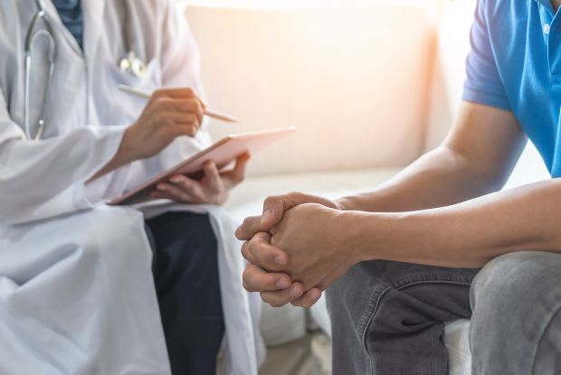 A doctor taking notes and a patient sitting with his hands clasped