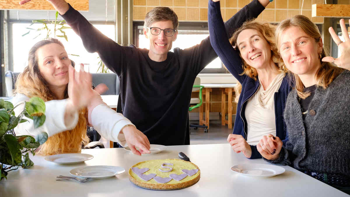 Das ForTomorrow-Team hat einen Kuchen auf dem Tisch. Eine lilane Ziffer fünf liegt auf dem Kuchen.