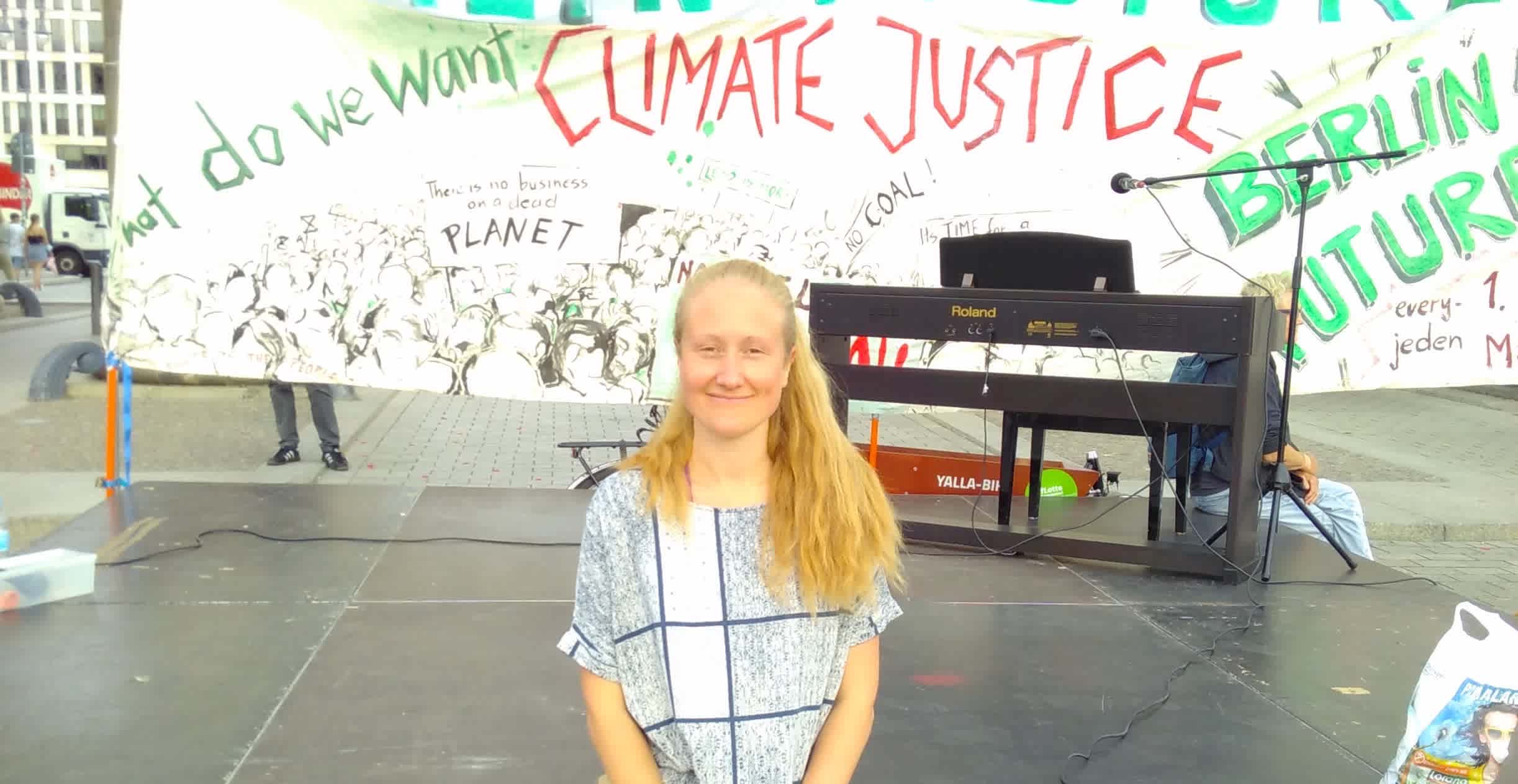 Jenny auf der Bühne auf einer Demo mit einem 'Climate Justice' Banner im Hintergrund