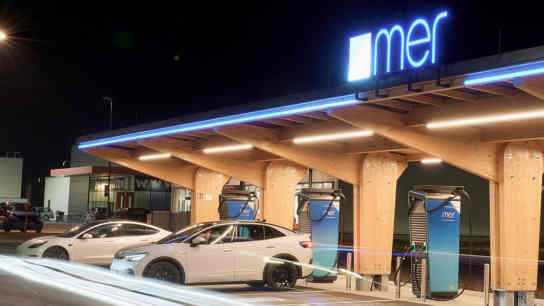 Vor einem Gebäude stehen Elektroautos, die an Mer-Ladestationen aufgeladen werden; im Hintergrund ist ein blaues Mer-Logo am Gebäude zu sehen