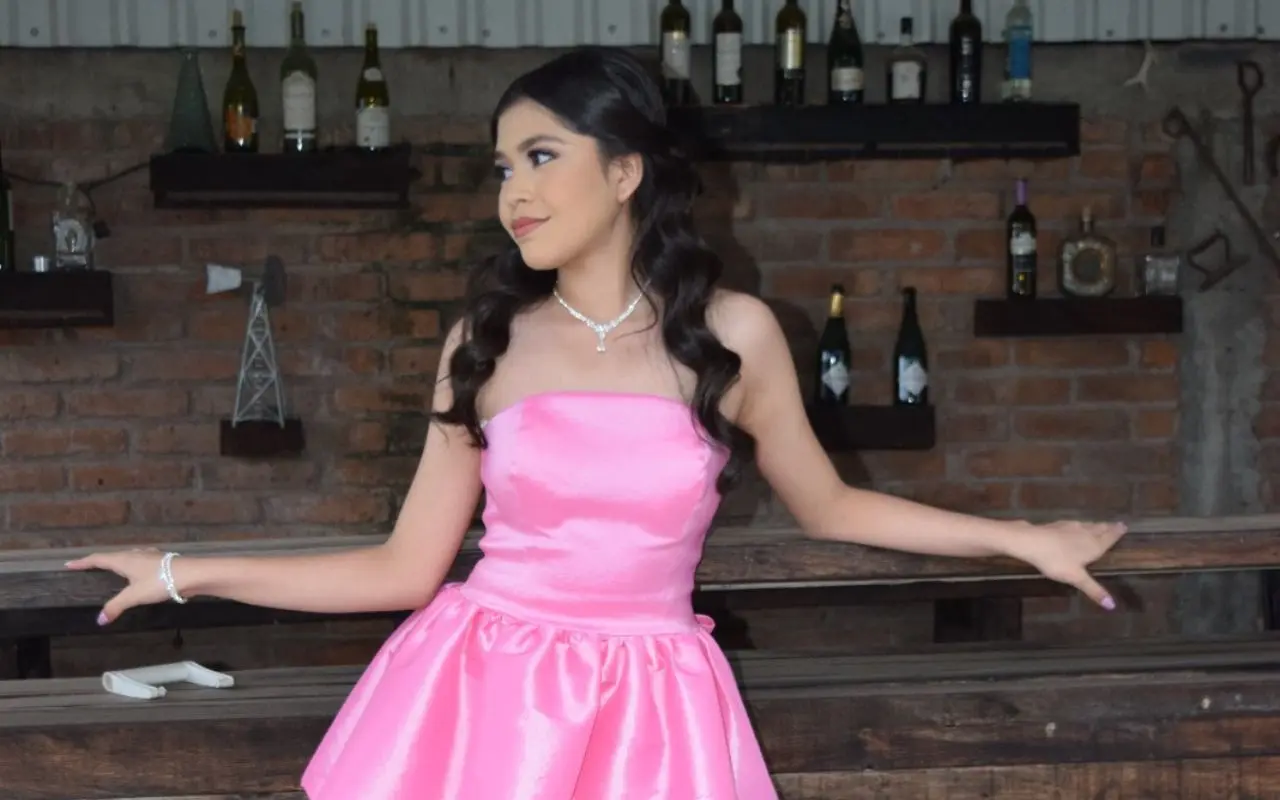 Estefanía Anguamea posando con vestido rosa satinado y accesorios elegantes, con un fondo rústico de madera.
