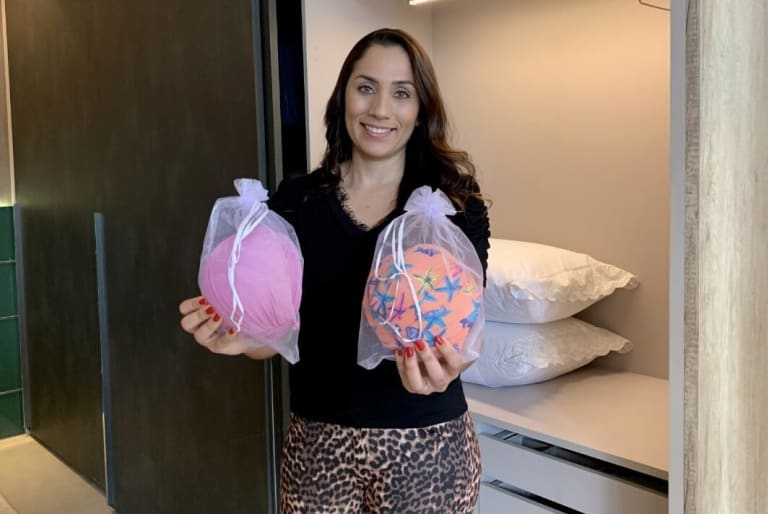 Há uma mulher no centro da foto segurando dois saquinhos organizadores com biquínis. Atrás dela, é possível ver um roupeiro na cor preta.