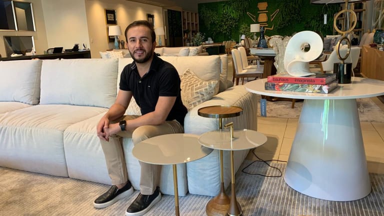 O designer está sentado em um sofá branco, com três mesinhas laterais ao seu lado.