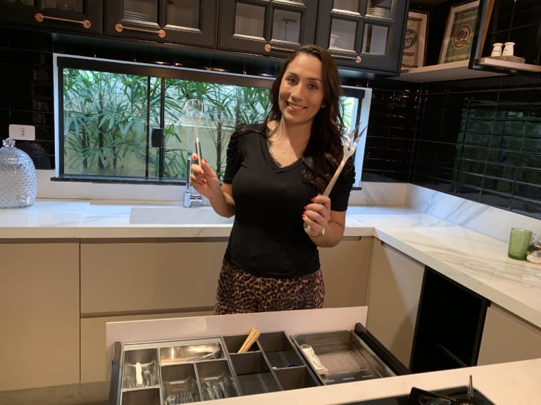 Na imagem, a personal organizer segura dois utensílios. Ao fundo, é possível ver uma cozinha preta com balcões brancos
