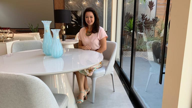 Na imagem, arquiteta está no centro, sentada em uma cadeira branca. Do lado esquerdo, é possível ver vasos de cristais longos na cor azul bebê.