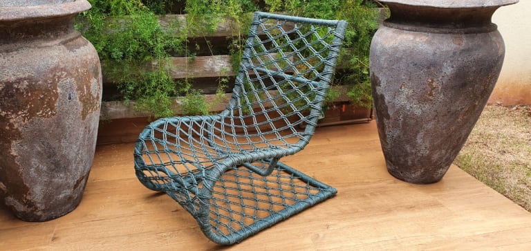 Na imagem, a poltrona Macros está no centro. Ao fundo, vê ripas de madeira e grandes vasos de decoração