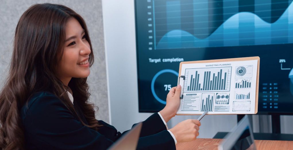 A businesswoman is holding a paper attached with a clipboard in one hand and with the other hand she is pointing to analytics data printed on that paper.