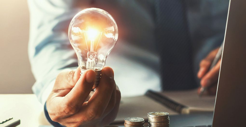 A businessman holding a light bulb that is glowing brightly upon two stacks of coins on the businessman's desk.