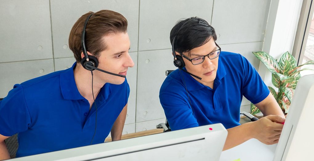 Two co-workers wearing blue T-shirts having a discussion about their works while working on their office desk.