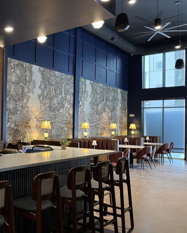Bar seating area with forest-toile wallpaper, navy panel walls, cane-back stools, and table lamps at Champagne Velvet Proper Kitchen & Bar