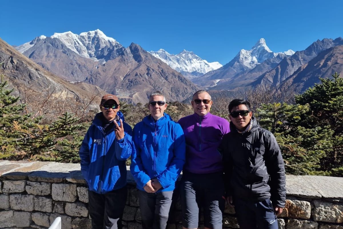 Four trekkers in blue jackets standing on a ridge above Namche with Everest and Ama Dablam on the horizon