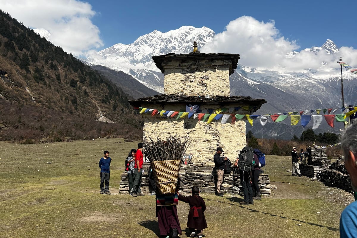 A whitewashed chorten draped in prayer flags on a grassy slope below snow peaks