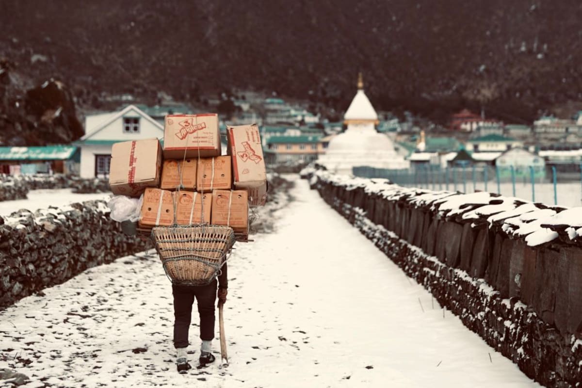 A porter carrying a tall load of supplies along a snowy path past a stupa in the Khumbu