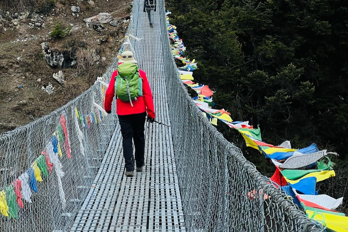 A trekker in a green jacket crossing a long suspension bridge strung with prayer flags in the Khumbu