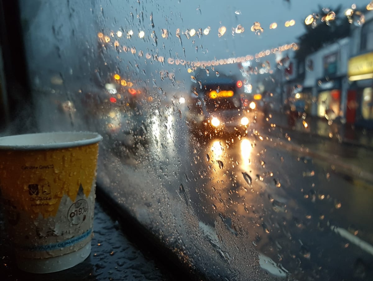 Rain-streaked window on a British morning commute, reflection of a commuter holding a takeaway coffee