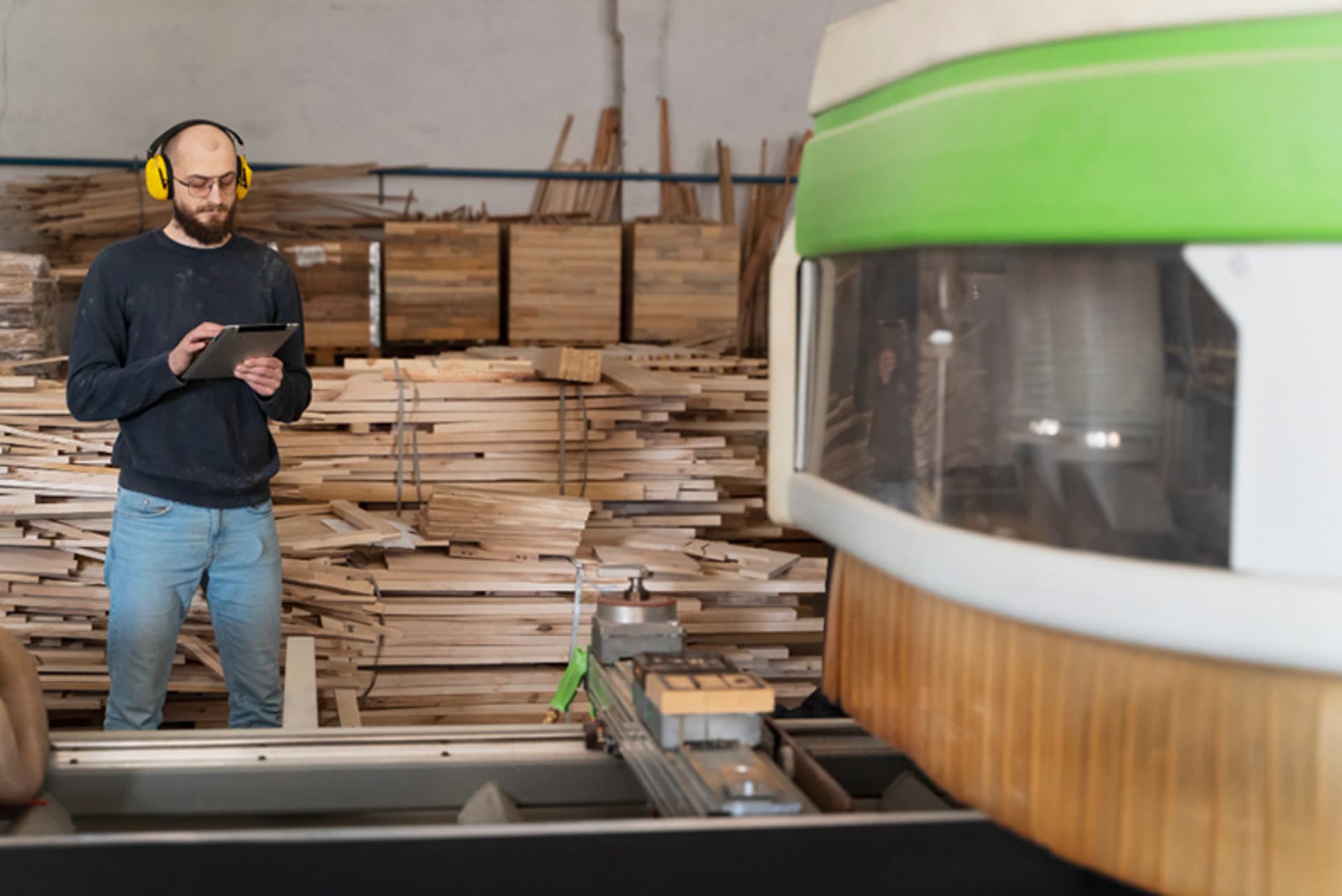 Holzbearbeitungsmaschinen in der Fertigung Mitarbeiter mit Tablet neben einer Holzbearbeitungsmaschine und gestapelten Holzbrettern in einer Werkstatt