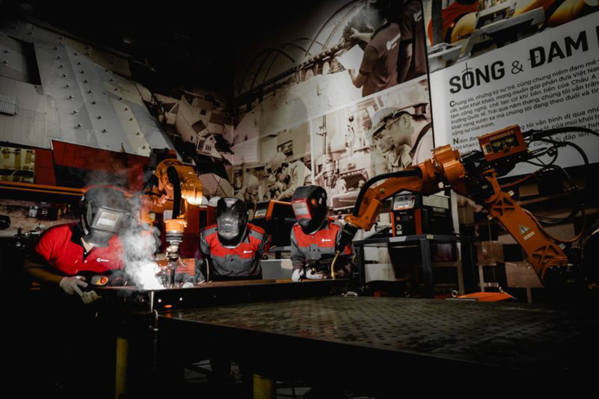 Three people wearing welding helmets at a welding table with orange robotic arms and a visible welding arc