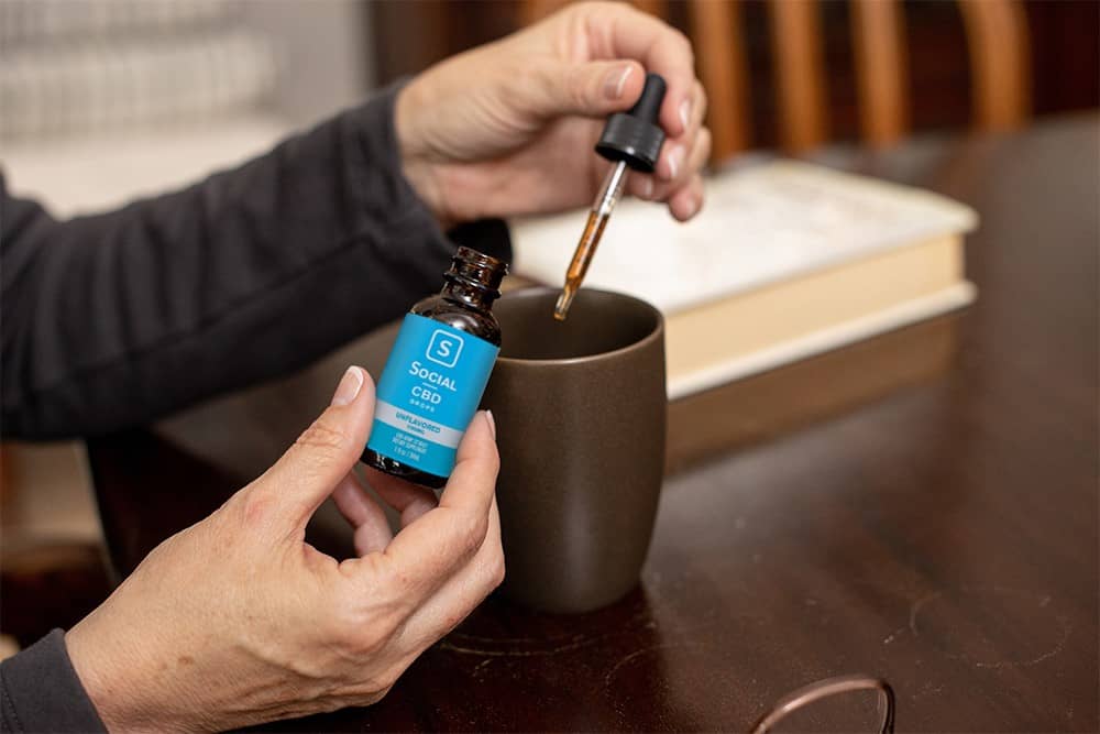 A user drops her dose of Social CBD tincture into a coffee mug while seated at a dark wood table