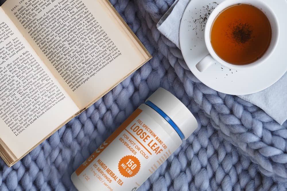 A bottle of CBD infused tea sits on a blue knit surface alongside a book and cup of tea