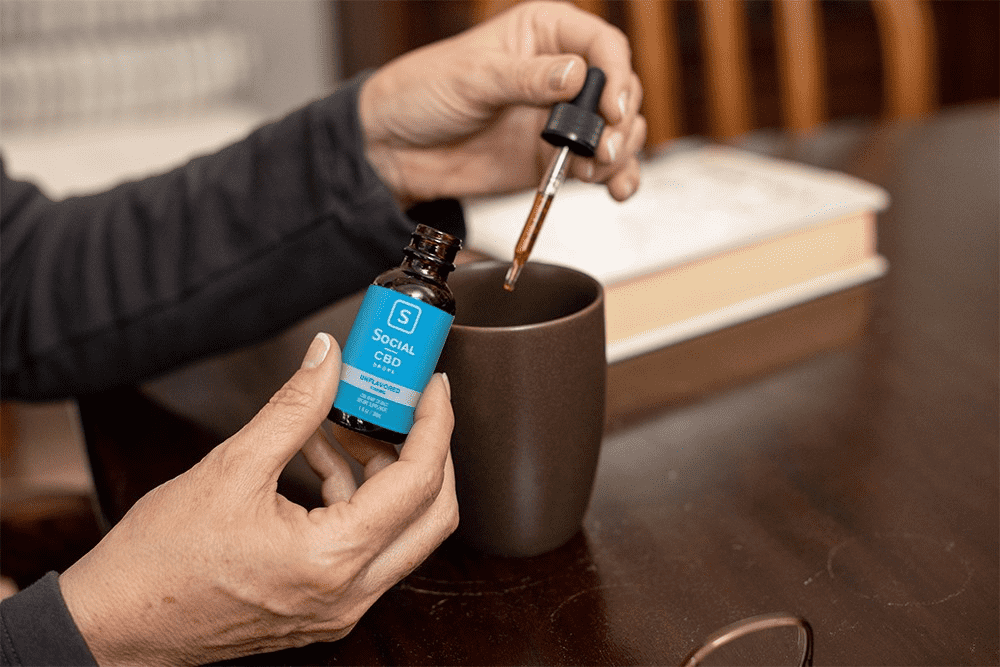 A user drops her dose of Social CBD tincture into a coffee mug while seated at a dark wood table