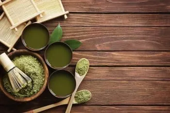 Two crossed wooden spoons and a wooden bowl filled with green powder, a bamboo whisk, and three cups of green liquid.