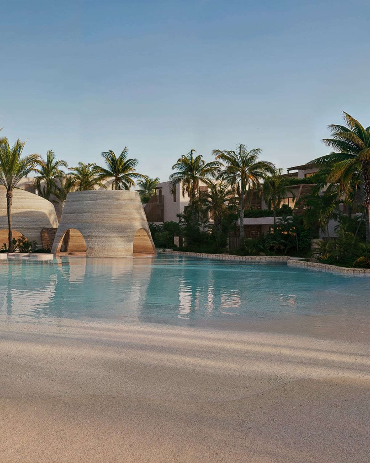 A lagoon pool with a sandy edge, woven cone-shaped pavilions and palm trees, Samaná Loft Villa · Render