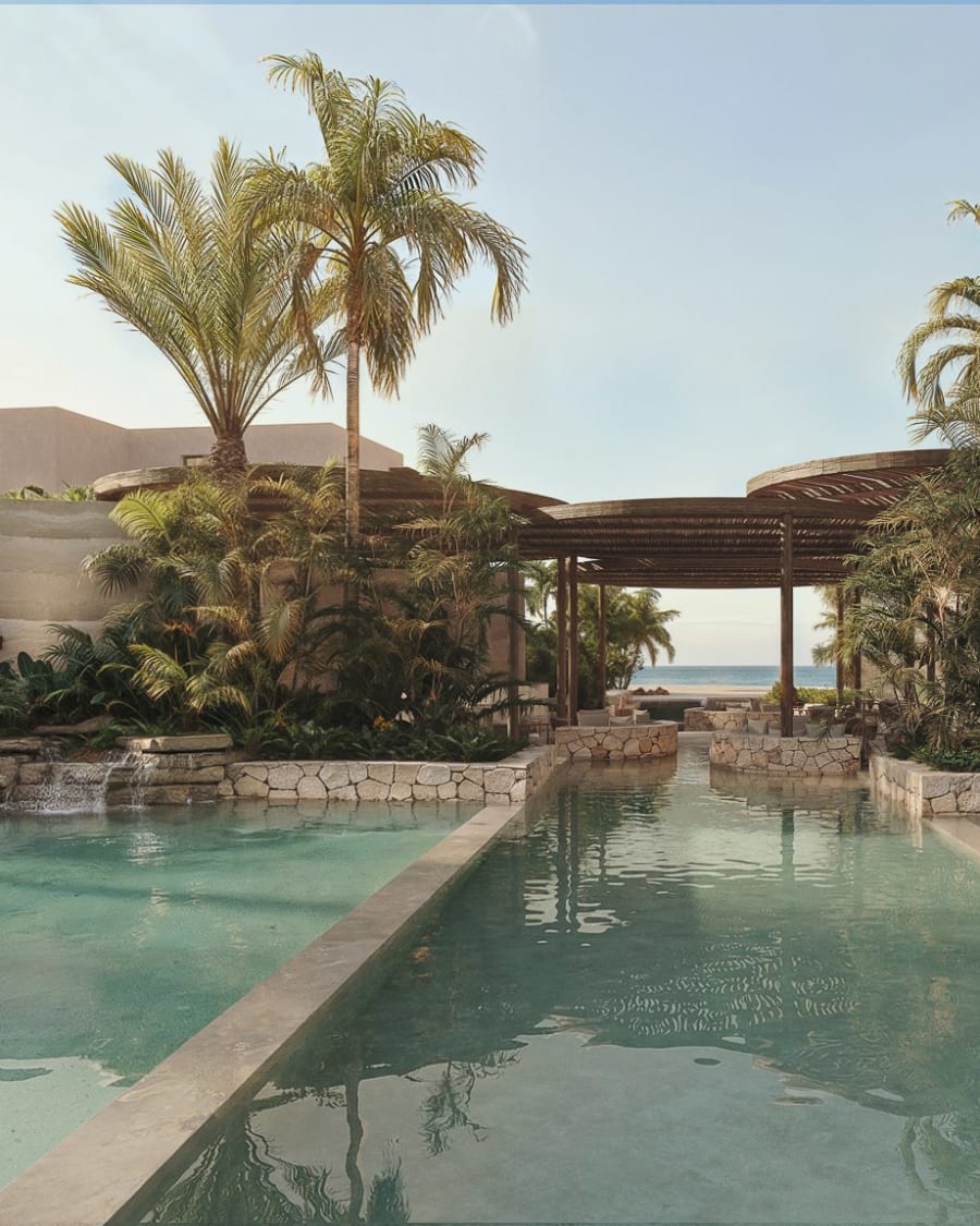A long pool between stone planters and palm trees, a pergola and the sea beyond