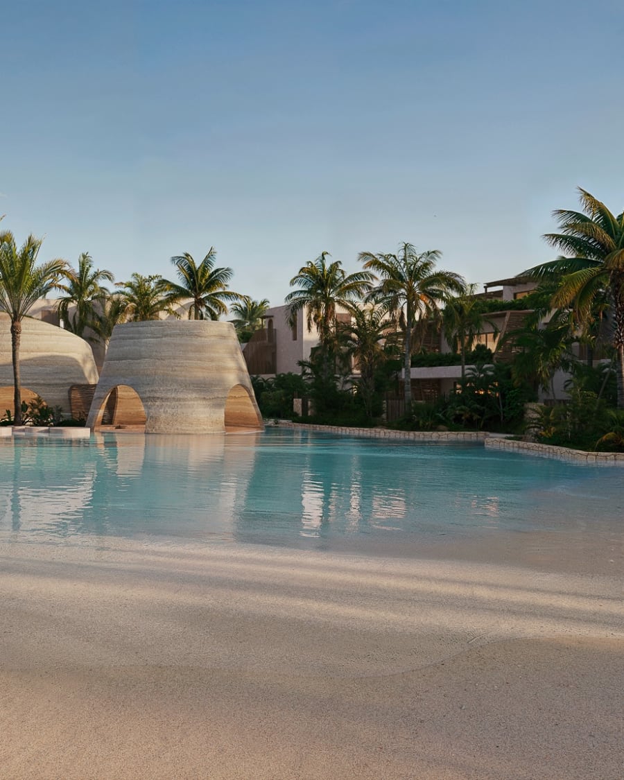 A lagoon pool with a sandy edge, woven cone-shaped pavilions and palm trees