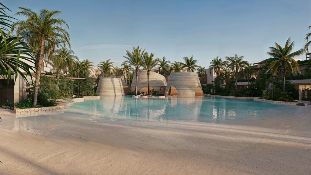 A lagoon pool with a sandy edge, woven cone-shaped pavilions and palm trees, Samaná Loft Villa · Render