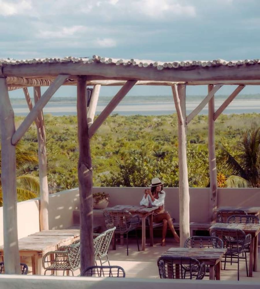 Unter einer Holzpergola blickt eine Terrasse über grünes Buschland auf eine Lagune und das Meer